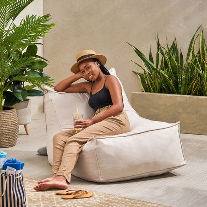 A woman in a hat, black tank top, and beige pants relaxes on the Ettie Water Resistant Fabric Bean Bag Chair. She holds a drink and smiles, surrounded by green plants in a bright, modern indoor space.