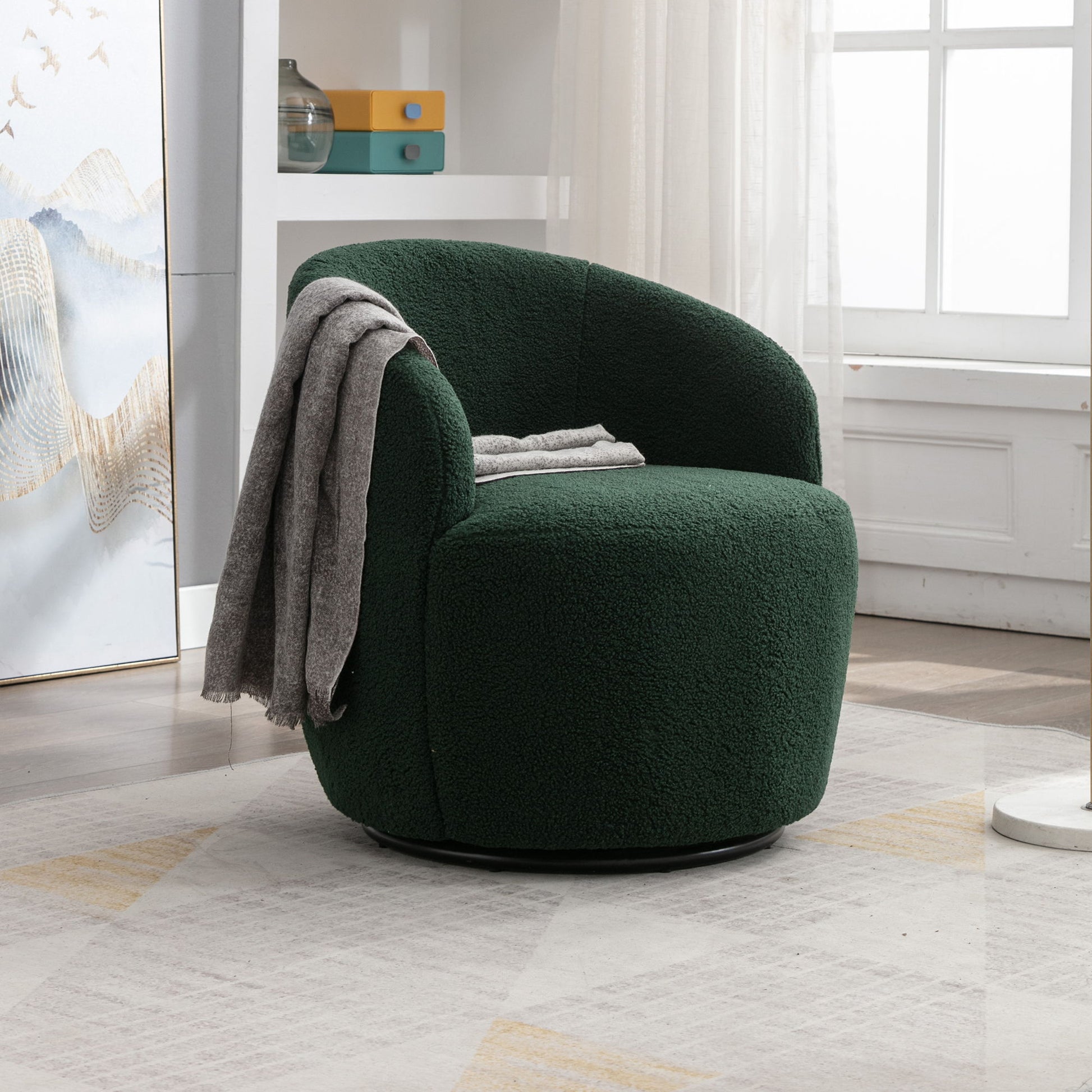 A Teddy Fabric Swivel Accent Armchair Barrel Chair with a powder coating metal ring, upholstered in dark green textured fabric, sits by a window in a bright room; a gray blanket rests on its curved backrest among modern decor.