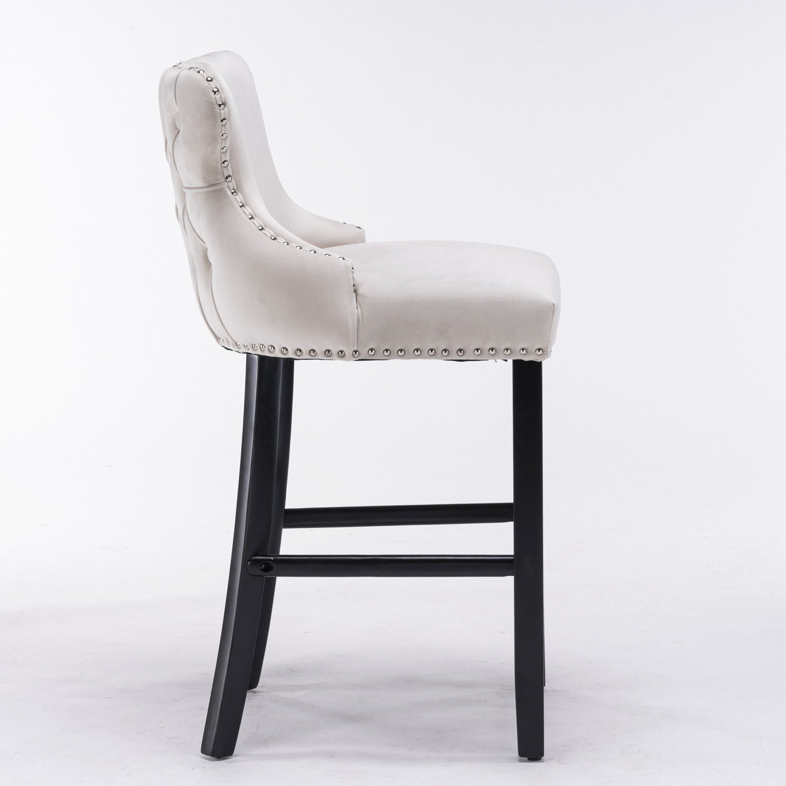 A side view of the Contemporary Velvet Upholstered Wing-Back Barstool set, featuring a light beige button-tufted seat and back, chrome nailhead trim, and wooden legs, shown against a plain white background.