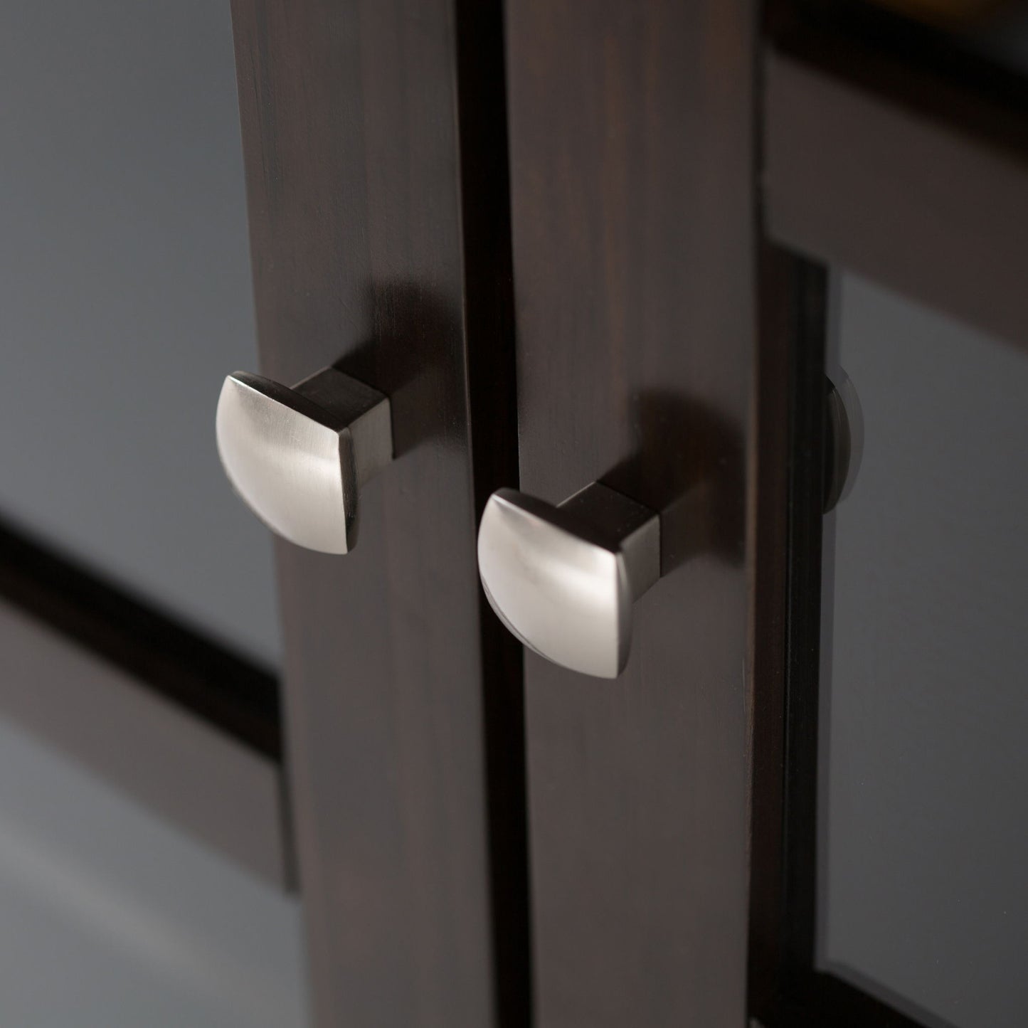 Close-up of two brushed metal knobs on the Acadian Low Storage Cabinet in Brunette Brown, with their reflection visible on the glass panel to the right.