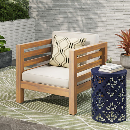 An Outdoor Acacia Wood Patio Chair With Fabric Cushion sits on a leaf-patterned rug by a navy blue side table with a cut-out design. Plants and a white brick wall are in the background, with white cushions and a patterned pillow on the chair.