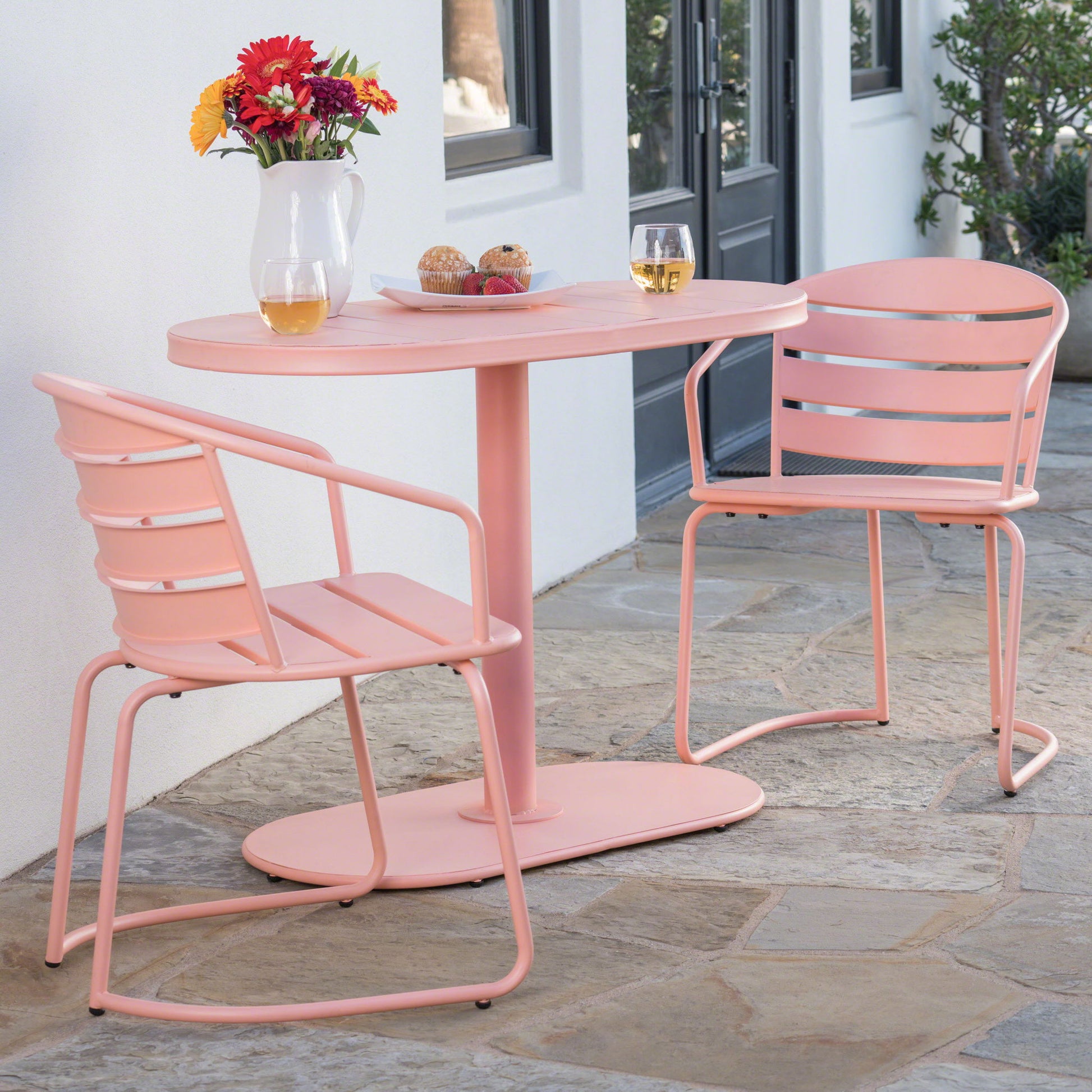 The 3 Piece Elegant Outdoor Bistro Set with 2 chairs and an oval table is arranged on a stone patio by a white wall and window, set for two with drinks, pastries, and a vase of flowers.