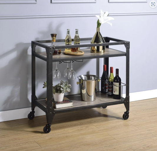 The Jorgensen Serving Cart in Rustic Oak and Charcoal holds liquor bottles, glasses, a gold vase with a white flower, a metal ice bucket, succulents, and wine bottles. It stands against a light gray wall on a wood floor.