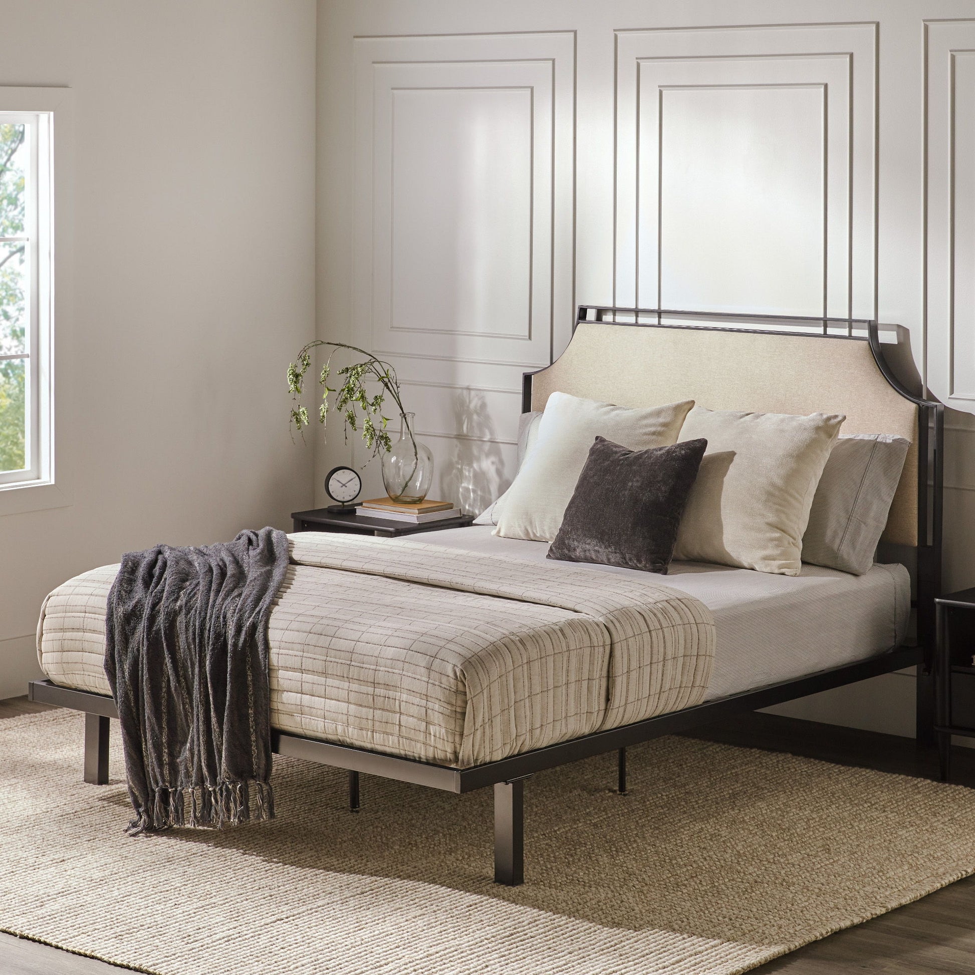 A modern bedroom featuring the Bed Traditional Upholstered Metal with cream and gray bedding, multiple pillows, a dark throw blanket, a bedside table with a clock, books, and plant, set against light paneled walls and a large window.