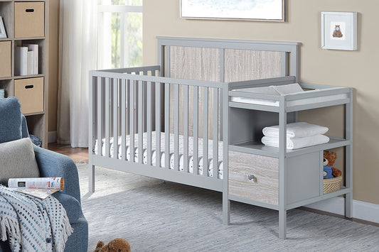 The Connelly 4-in-1 Crib and Changer Combo, in light gray with shelves and a drawer, is set in a cozy nursery featuring a blue armchair, gray rug, soft blankets, and nearby window with wall art.