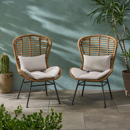 Two Calypso Outdoor Chairs with water-resistant beige cushions and iron frames sit side by side on a tiled patio, surrounded by potted plants—a cactus and a tall leafy variety—set against a green textured wall.