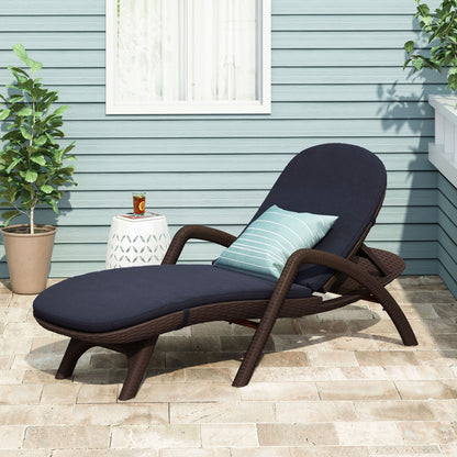 The Mikael - Chaise Lounge With Cushion in brown wicker features a navy cushion and a striped pillow on a stone patio, alongside two potted plants and a small white side table with iced tea; a light blue house stands in the background.