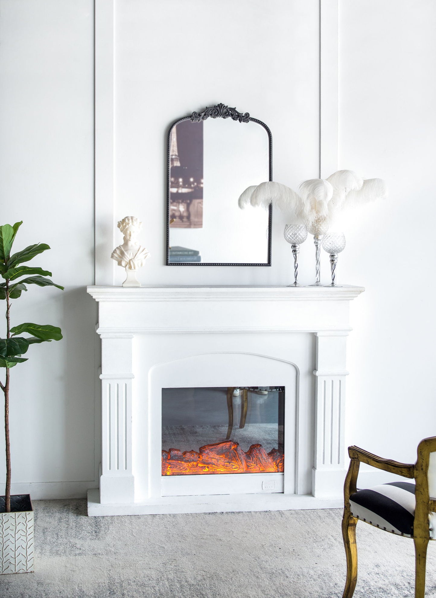 A white fireplace with a glowing electric fire sits against a white wall. Above it hangs the Arch Mirror, Baroque Inspired Wall Decor, reflecting part of a city photo. Decor includes a plant, bust, feather arrangement, and gold-accented chair.