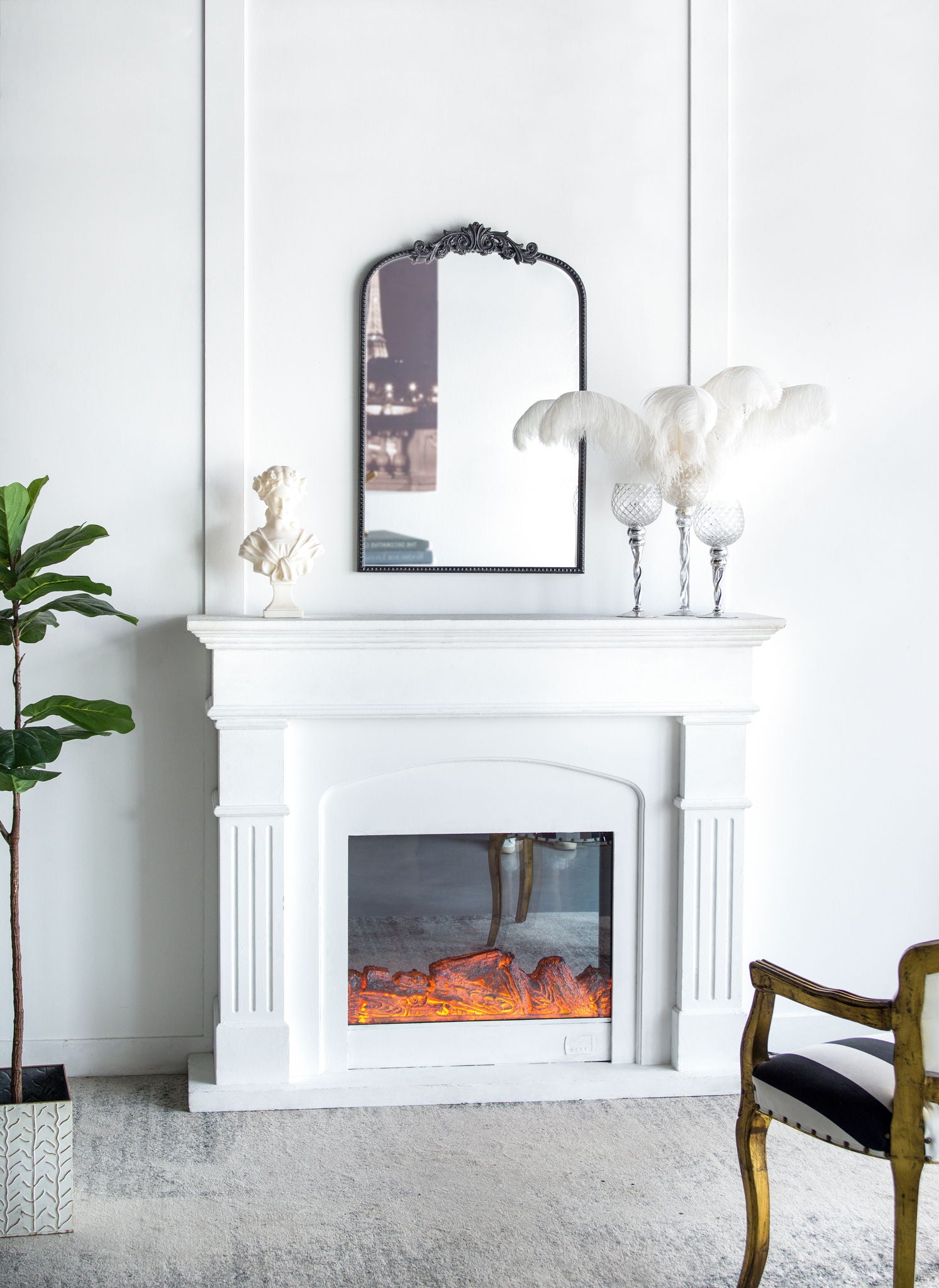 A white fireplace with a glowing electric fire sits against a white wall. Above it hangs the Arch Mirror, Baroque Inspired Wall Decor, reflecting part of a city photo. Decor includes a plant, bust, feather arrangement, and gold-accented chair.
