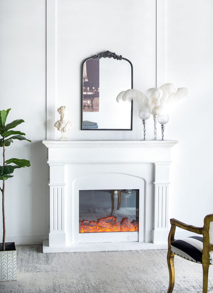 A white fireplace with a glowing electric fire sits against a white wall. Above it hangs the Arch Mirror, Baroque Inspired Wall Decor, reflecting part of a city photo. Decor includes a plant, bust, feather arrangement, and gold-accented chair.