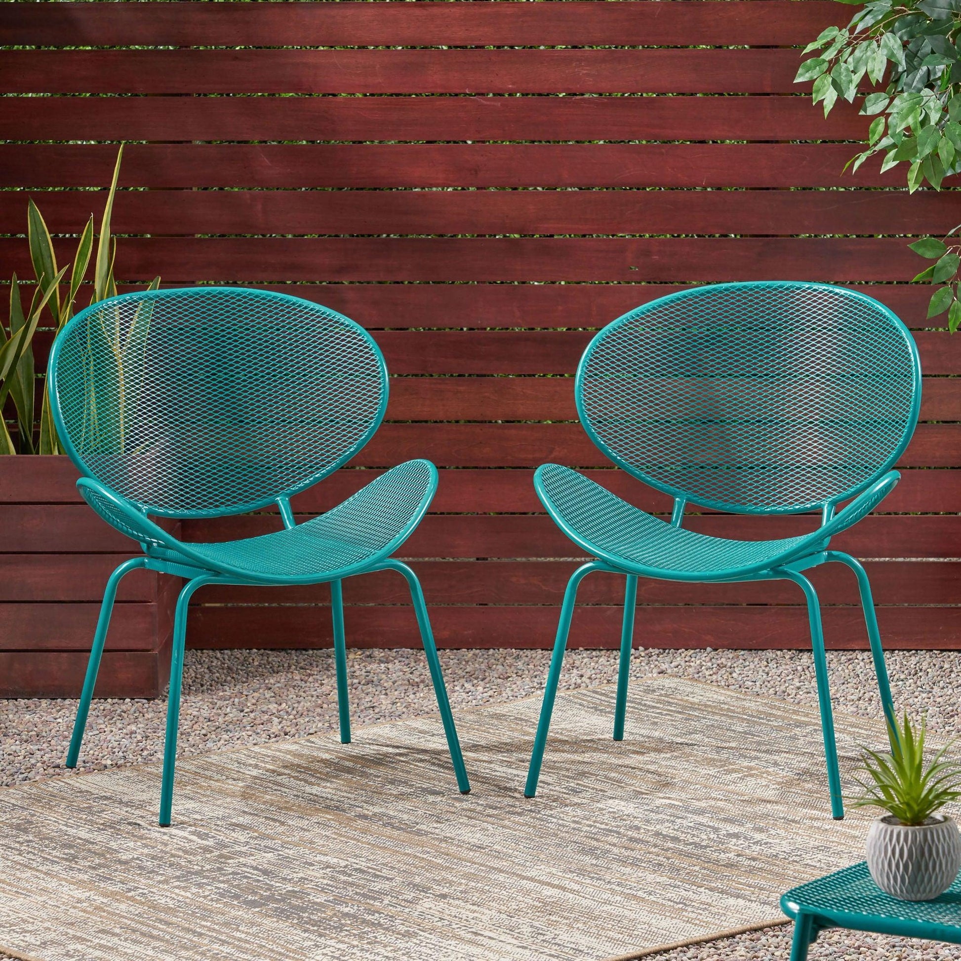 The Elba - Contemporary Chair Set For Outdoor Relaxation features two teal metal chairs with mesh seats and backs, placed on a textured rug amid potted plants, gravel, and a wooden slat fence with lush green foliage behind.