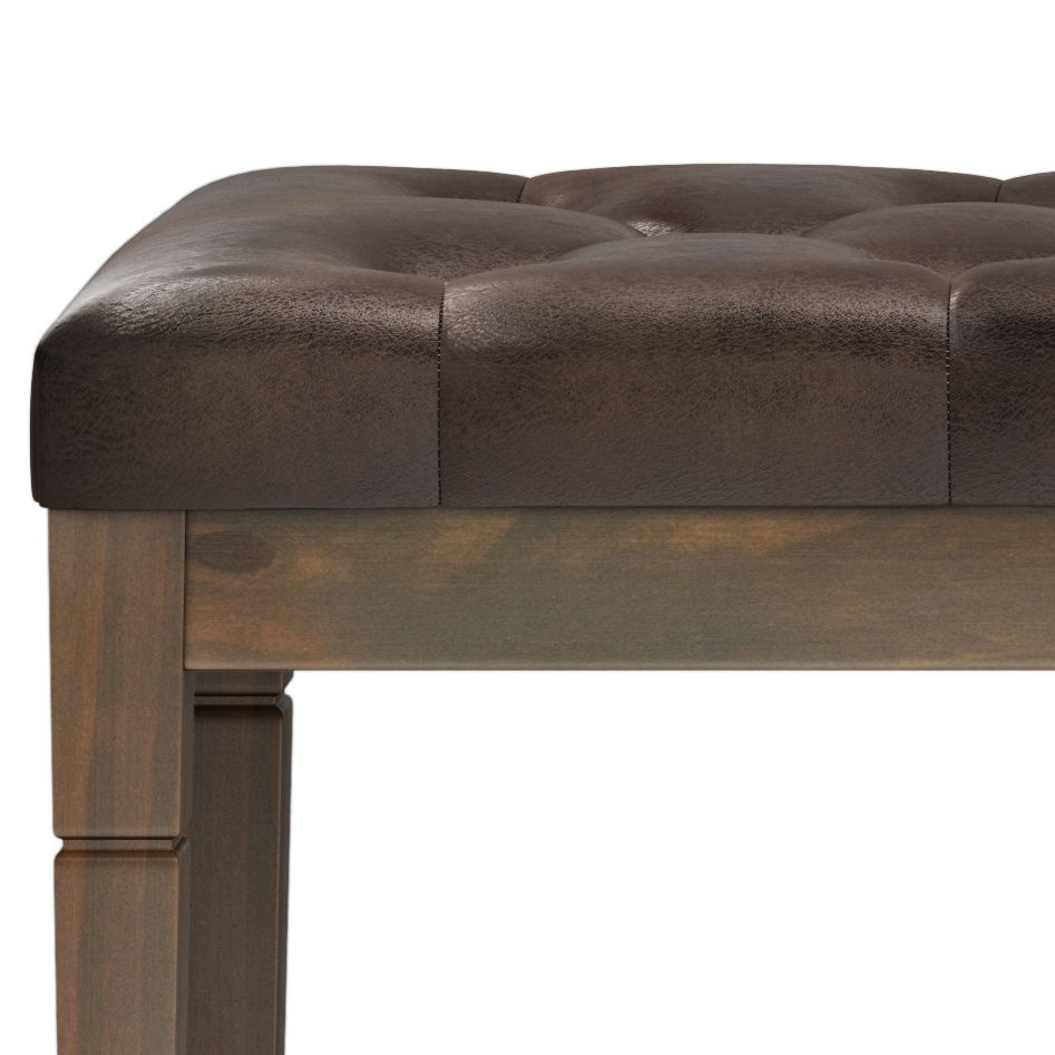 Close-up of the Waverly - Upholstered Tufted Ottoman Bench featuring a dark brown, tufted leather seat, square edges, and carved wooden legs.