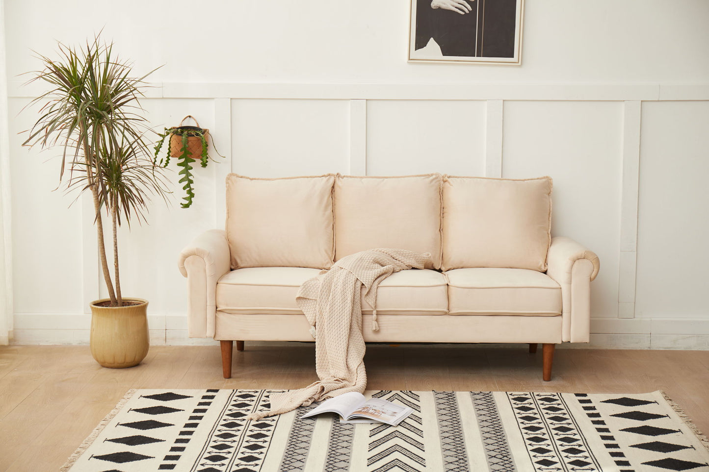 The Graceful Design Plush Comfort Sofa, in beige, sits by a white paneled wall with a light knit blanket over its arm. A potted plant is on the left, a hanging plant decorates the wall, and a geometric rug with an open book lies on the floor.