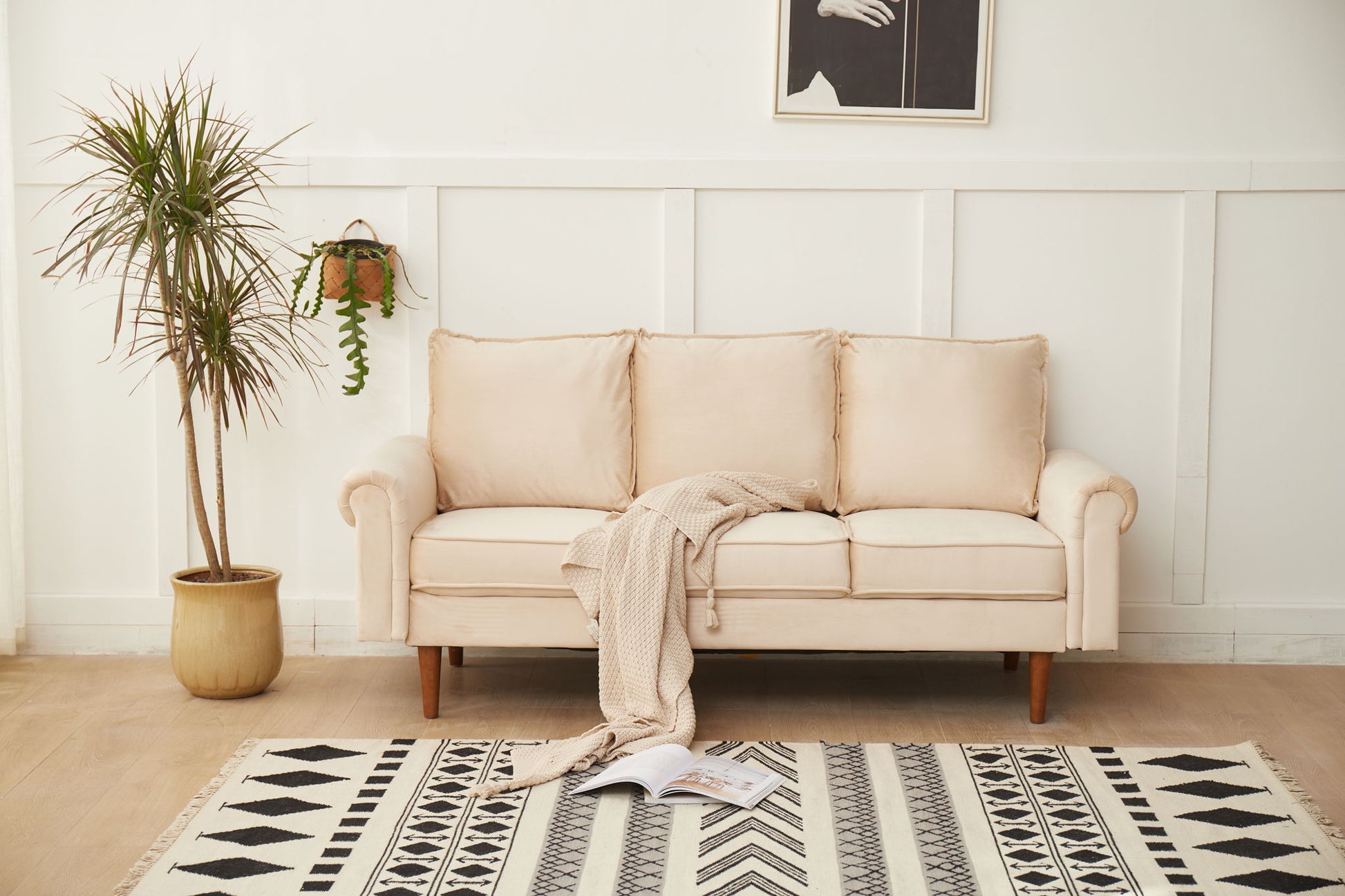 The Graceful Design Plush Comfort Sofa, in beige, sits by a white paneled wall with a light knit blanket over its arm. A potted plant is on the left, a hanging plant decorates the wall, and a geometric rug with an open book lies on the floor.