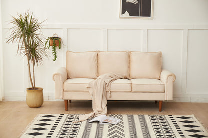 The Graceful Design Plush Comfort Sofa, in beige, sits by a white paneled wall with a light knit blanket over its arm. A potted plant is on the left, a hanging plant decorates the wall, and a geometric rug with an open book lies on the floor.