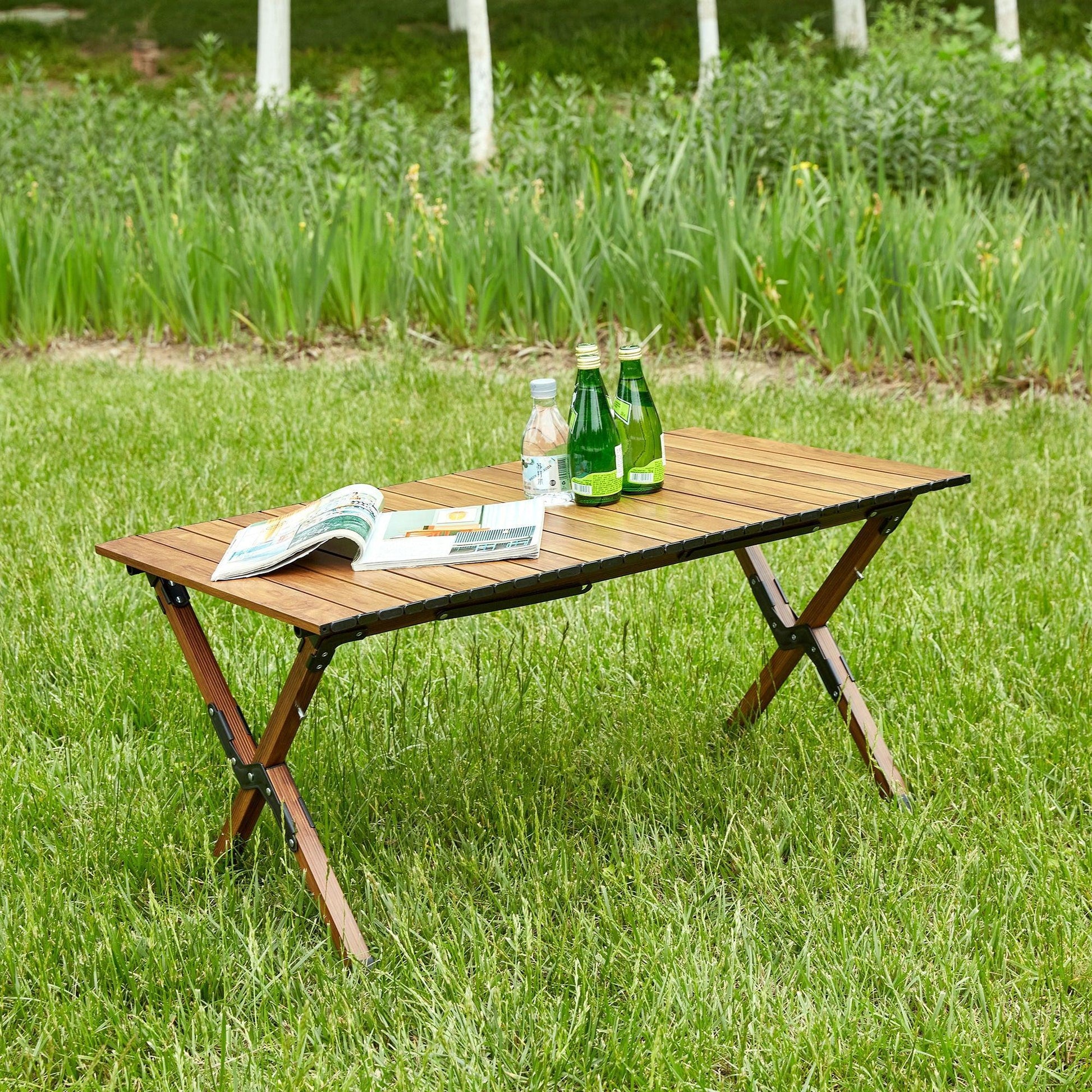 A Folding Outdoor Table (Lightweight Aluminum Roll-Up, Brown) stands on green grass, holding an open book, a notebook, two sparkling water bottles, and two water bottles, with trees and tall grass in the background.