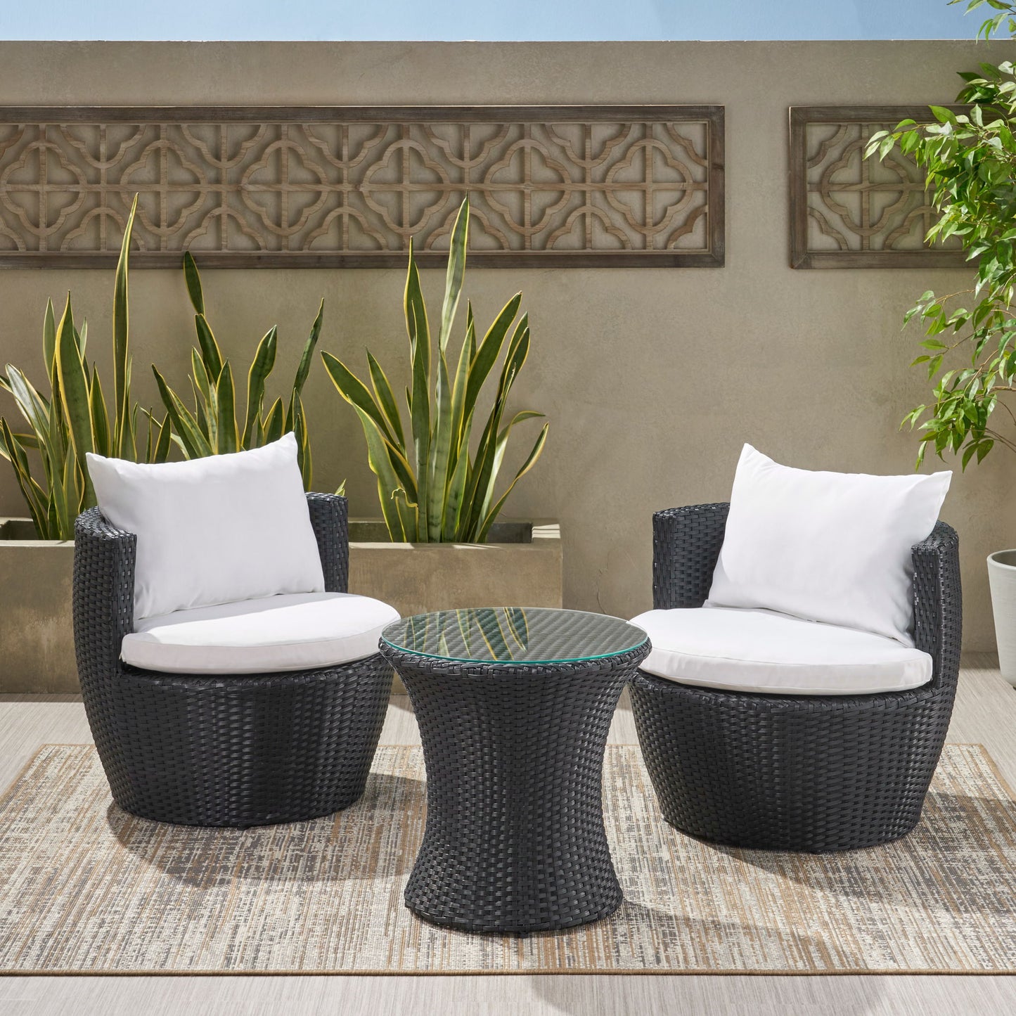 The Kono Chat Set Outdoor Furniture features two black wicker chairs with white cushions and a matching round table, arranged on an outdoor rug with tall green plants and a decorative concrete wall in the background.