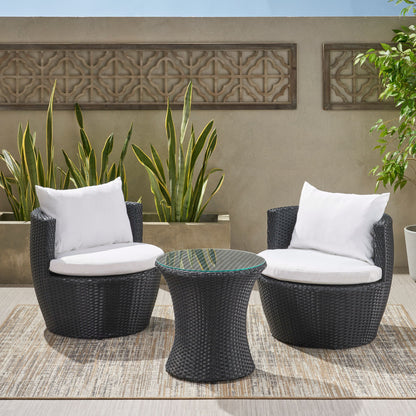 The Kono Chat Set Outdoor Furniture features two black wicker chairs with white cushions and a matching round table, arranged on an outdoor rug with tall green plants and a decorative concrete wall in the background.