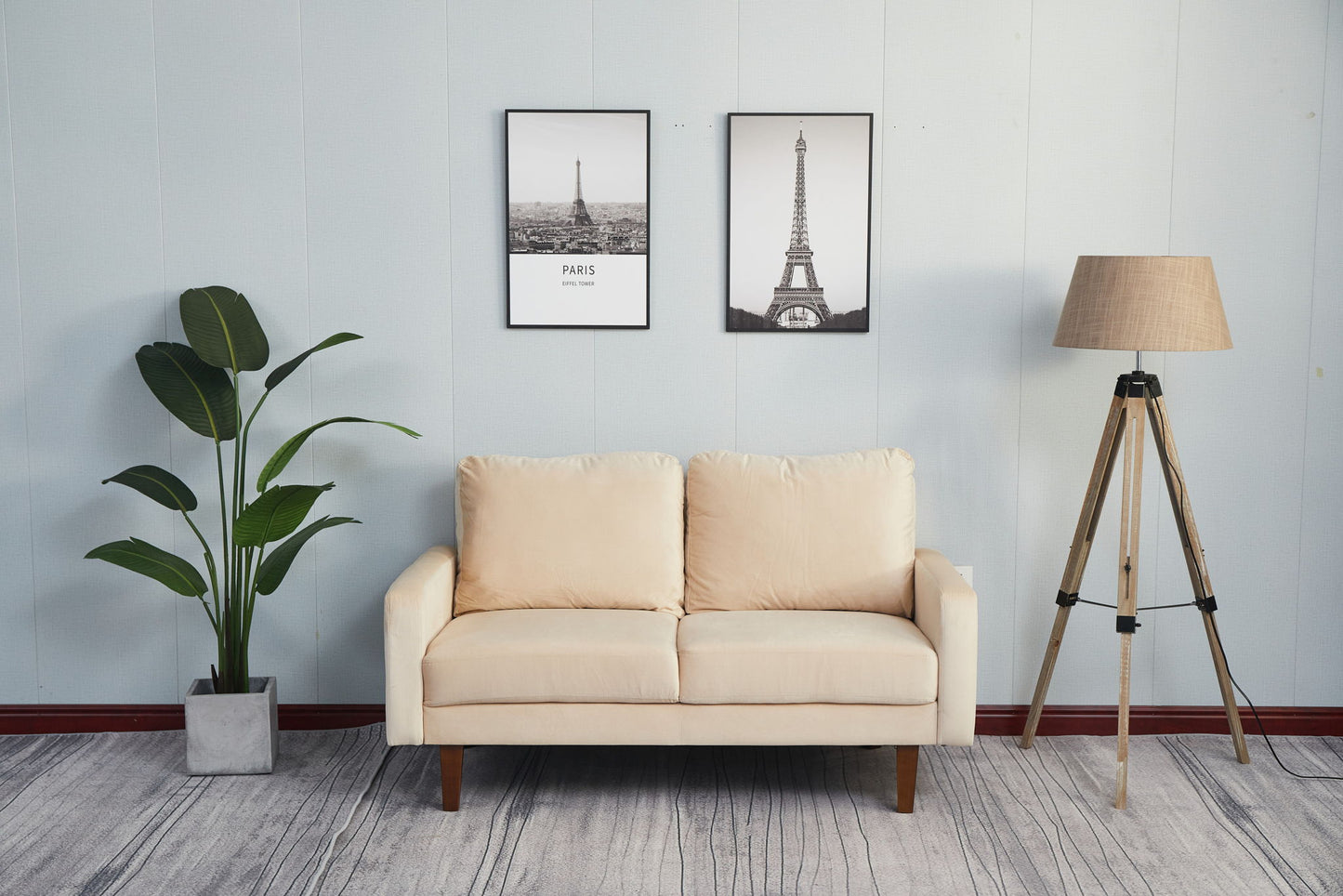 The Comfortable Sofa With Durable Construction is positioned against a light blue wall with two framed pictures above, flanked by a potted plant on the left and a wooden floor lamp with a beige shade on the right. The floor features a gray pattern.