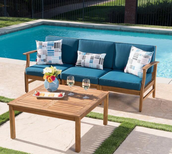 The Perla - 3 Seater Sofa And Table Set with blue cushions and patterned pillows sits poolside, paired with a wooden table holding white wine, flowers, and fruit. Green grass and a fence complete the outdoor scene.
