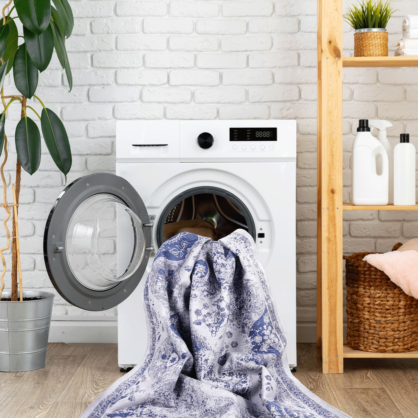 A Maval - Stunning Area Rug is draped from the open door of a front-loading washing machine; nearby are a potted plant and a wooden shelf stocked with towels and laundry supplies.