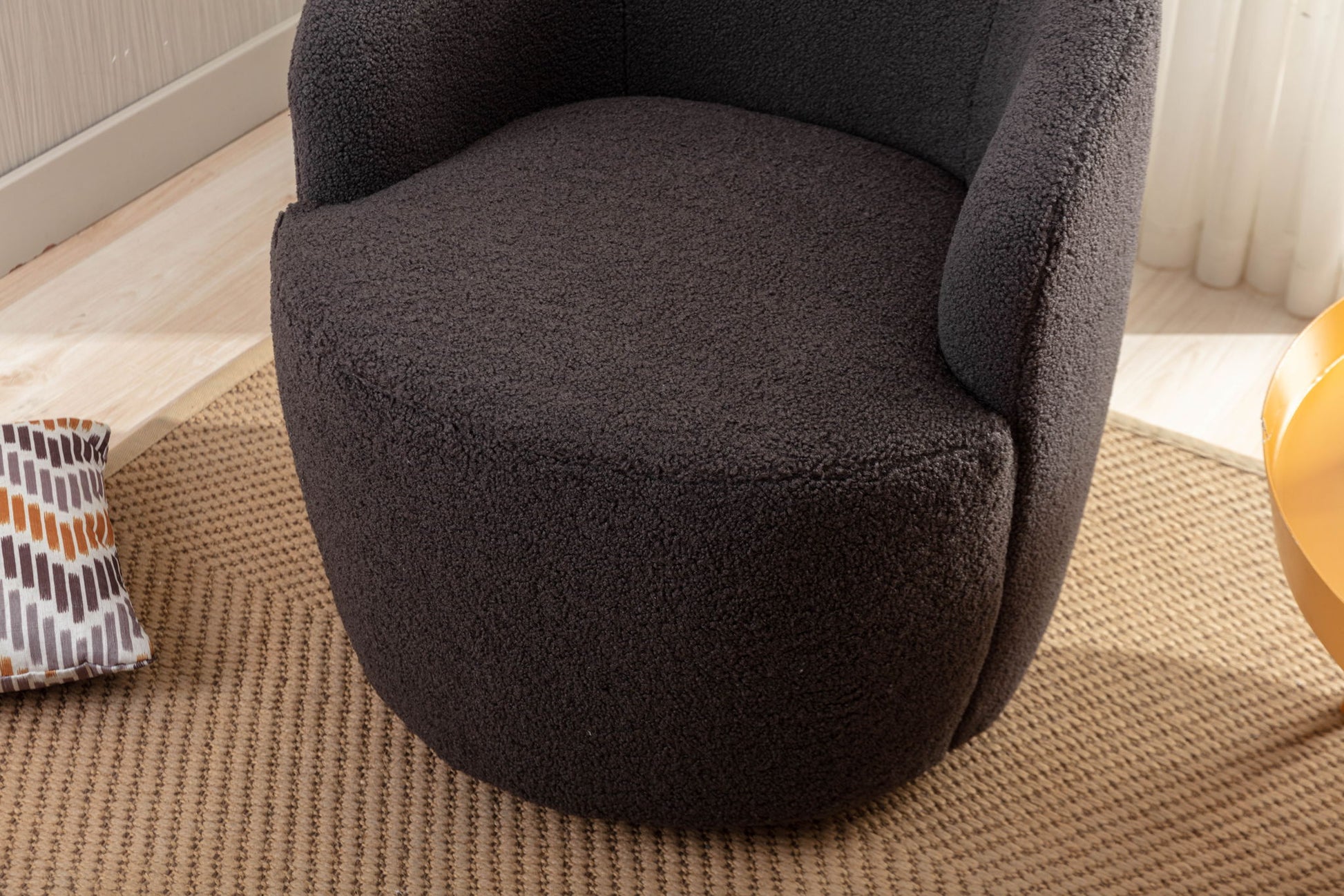 A close-up of the Teddy Fabric Swivel Accent Armchair Barrel Chair with powder-coated metal ring in dark gray, set on a textured beige rug, partially showing a patterned cushion and a nearby yellow object.