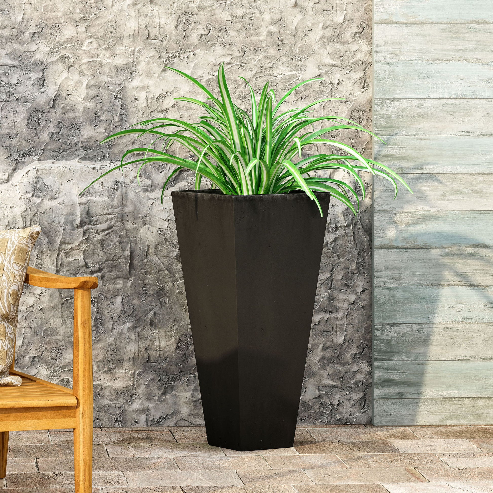 An Outdoor Modern Cast Stone Planter in black holds a spider plant with green and white leaves, placed on a stone patio by a textured gray wall, with a wooden bench partially visible on the left.