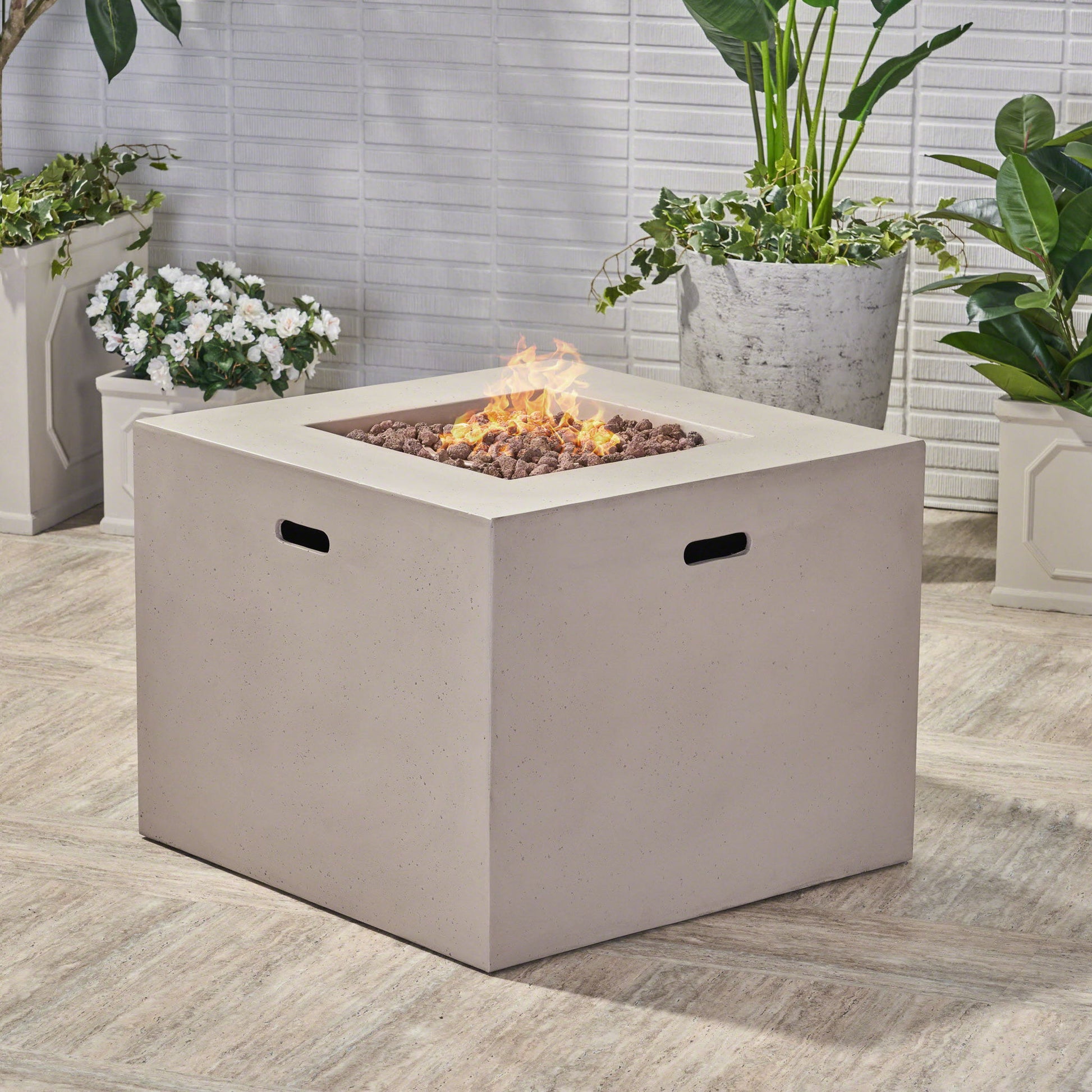 The Outdoor Patio Square Gas Burning Concrete Fire Pit 40,000 Btu, featuring a modern gray finish and lively flames, is surrounded by potted green plants and white flowers on a tiled patio.