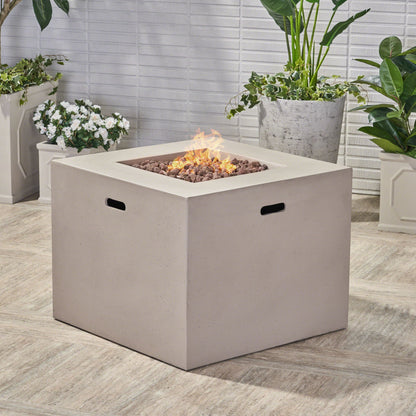 The Outdoor Patio Square Gas Burning Concrete Fire Pit 40,000 Btu, featuring a modern gray finish and lively flames, is surrounded by potted green plants and white flowers on a tiled patio.