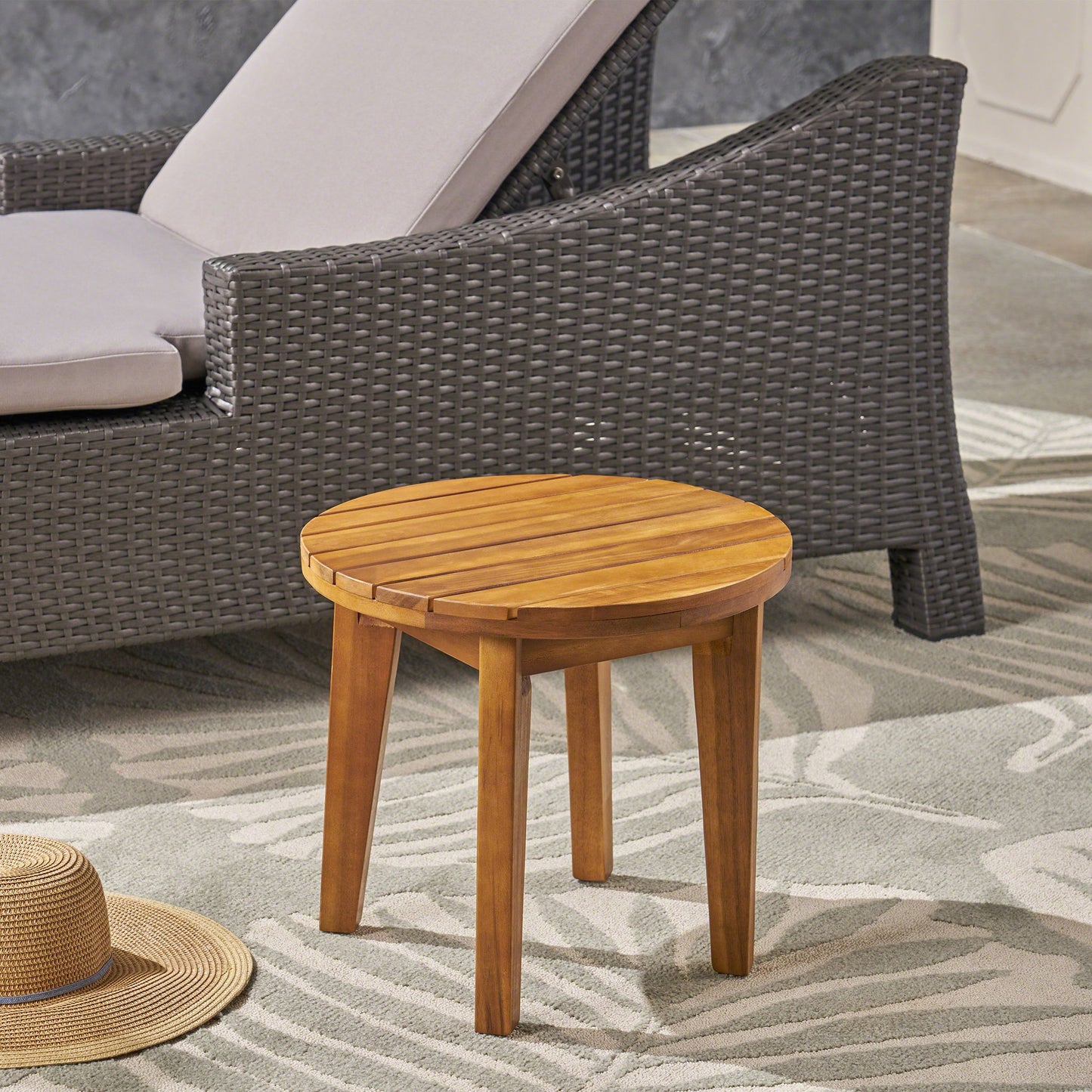 An Outdoor Acacia Wood Side Table With Weather Resistance stands on a patterned rug beside a wicker lounge chair with light cushions, while a straw hat rests on the floor nearby.