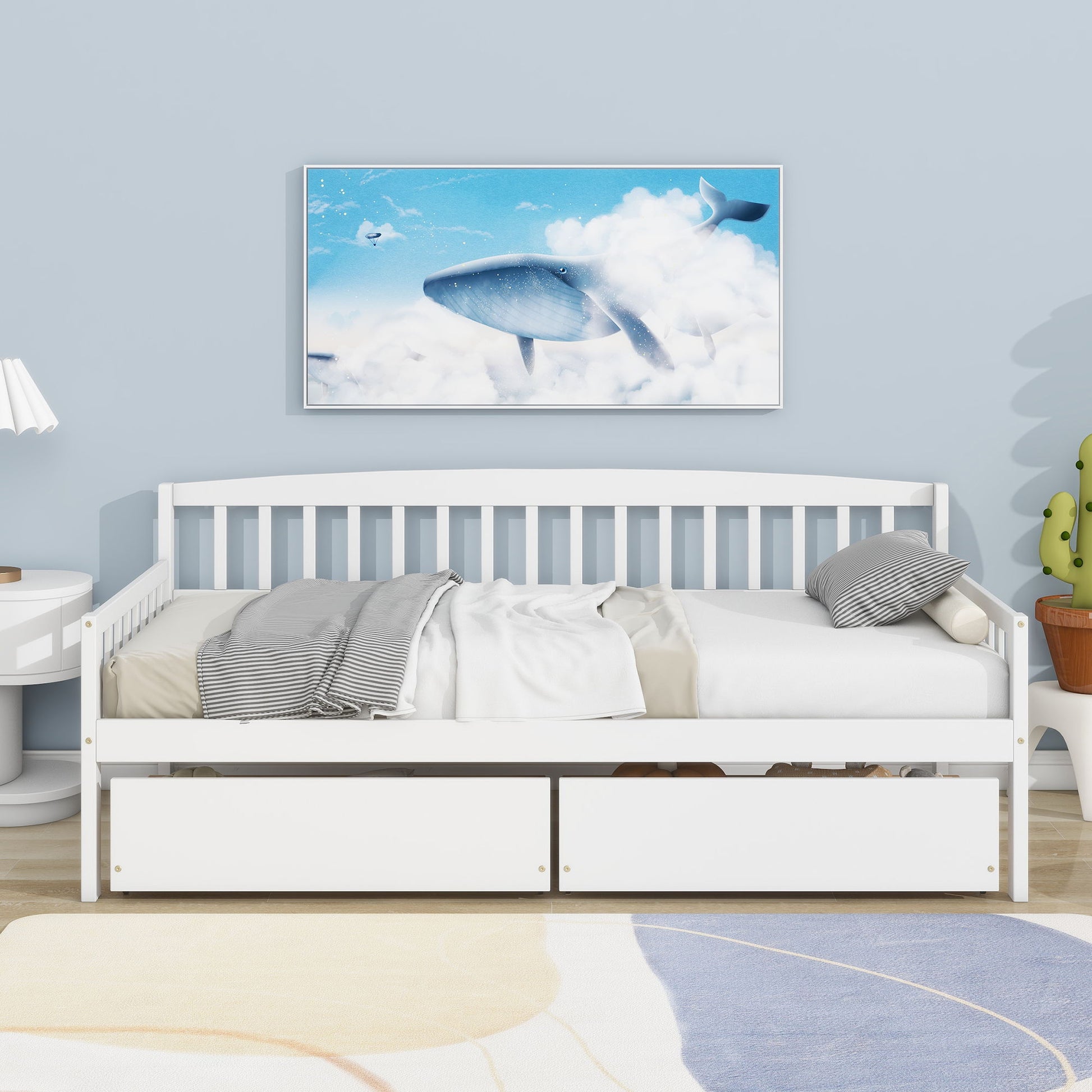 A Pine Wood Daybed with two storage drawers and striped bedding rests against a light blue wall; above is a framed whale-and-cloud print, while a cactus-shaped décor and lamp are placed nearby.
