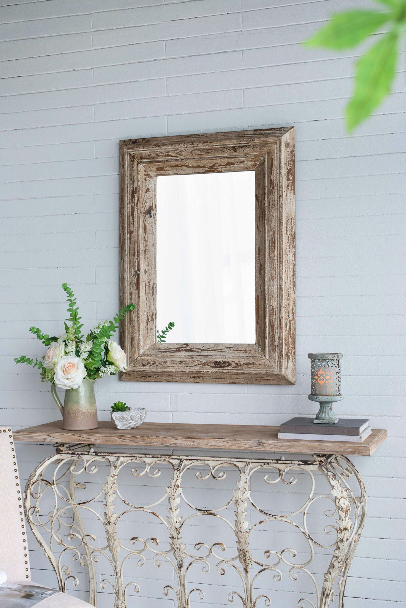 A Rectangle Wall Accent Mirror With Distressed Wood Frame - Brown hangs on a light gray brick wall above a vintage console table, which displays a vase with white flowers, greenery, a candle, and a small decorative dish.