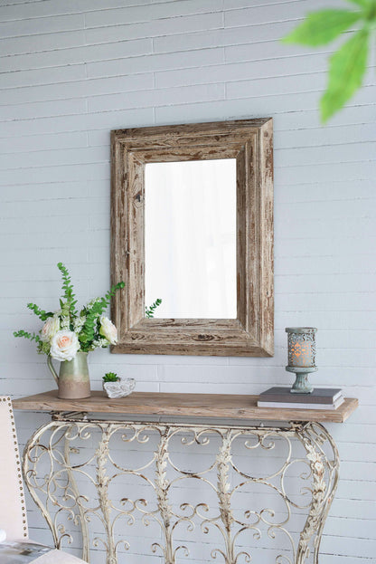 A Rectangle Wall Accent Mirror With Distressed Wood Frame - Brown hangs on a light gray brick wall above a vintage console table, which displays a vase with white flowers, greenery, a candle, and a small decorative dish.