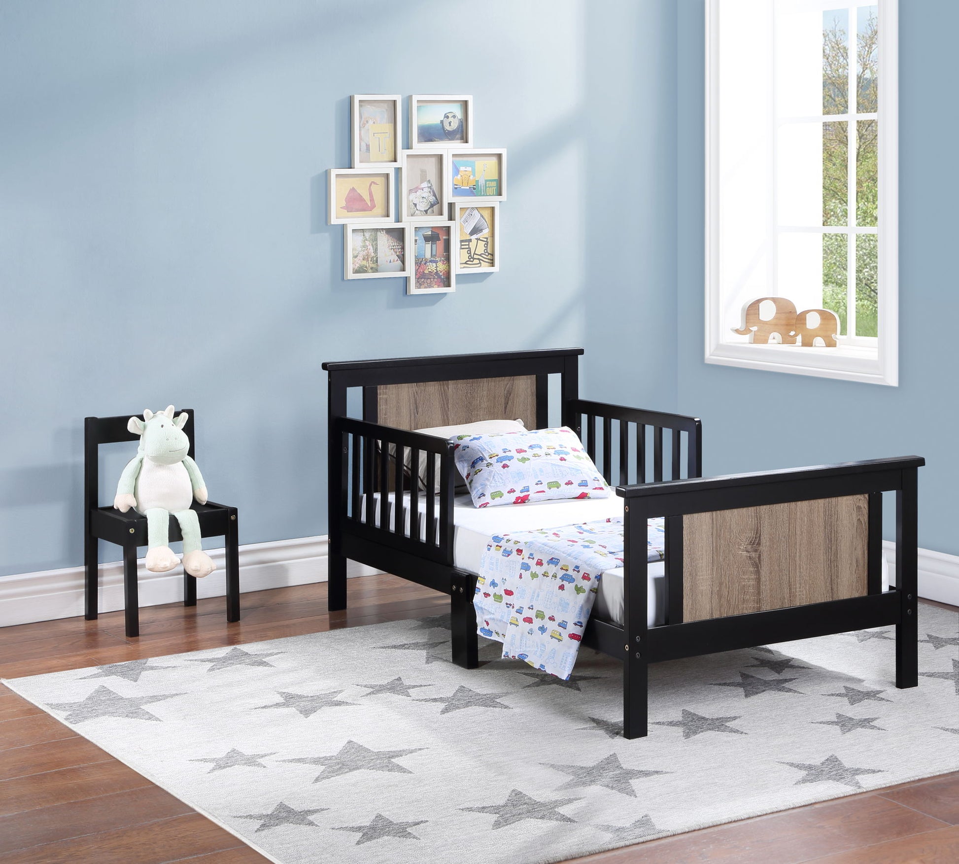 A Connelly Reversible Panel Toddler Bed with black frame and wood panels sits in a light blue room, paired with a small chair holding a stuffed unicorn, a gray star rug, wall art, and sunlight from the window for a cozy, kid-friendly space.