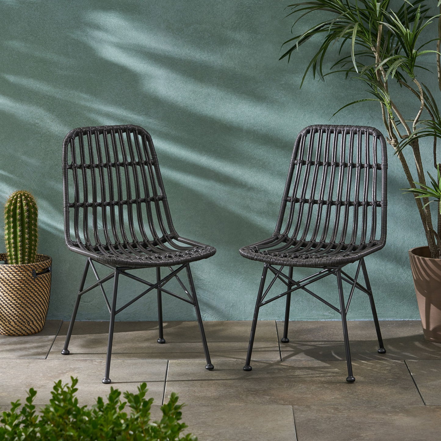Two Sawtelle - Chairs in black woven rattan with metal legs sit side by side on tiled flooring, flanked by potted plants such as a cactus and tall palm, set against a green textured wall.