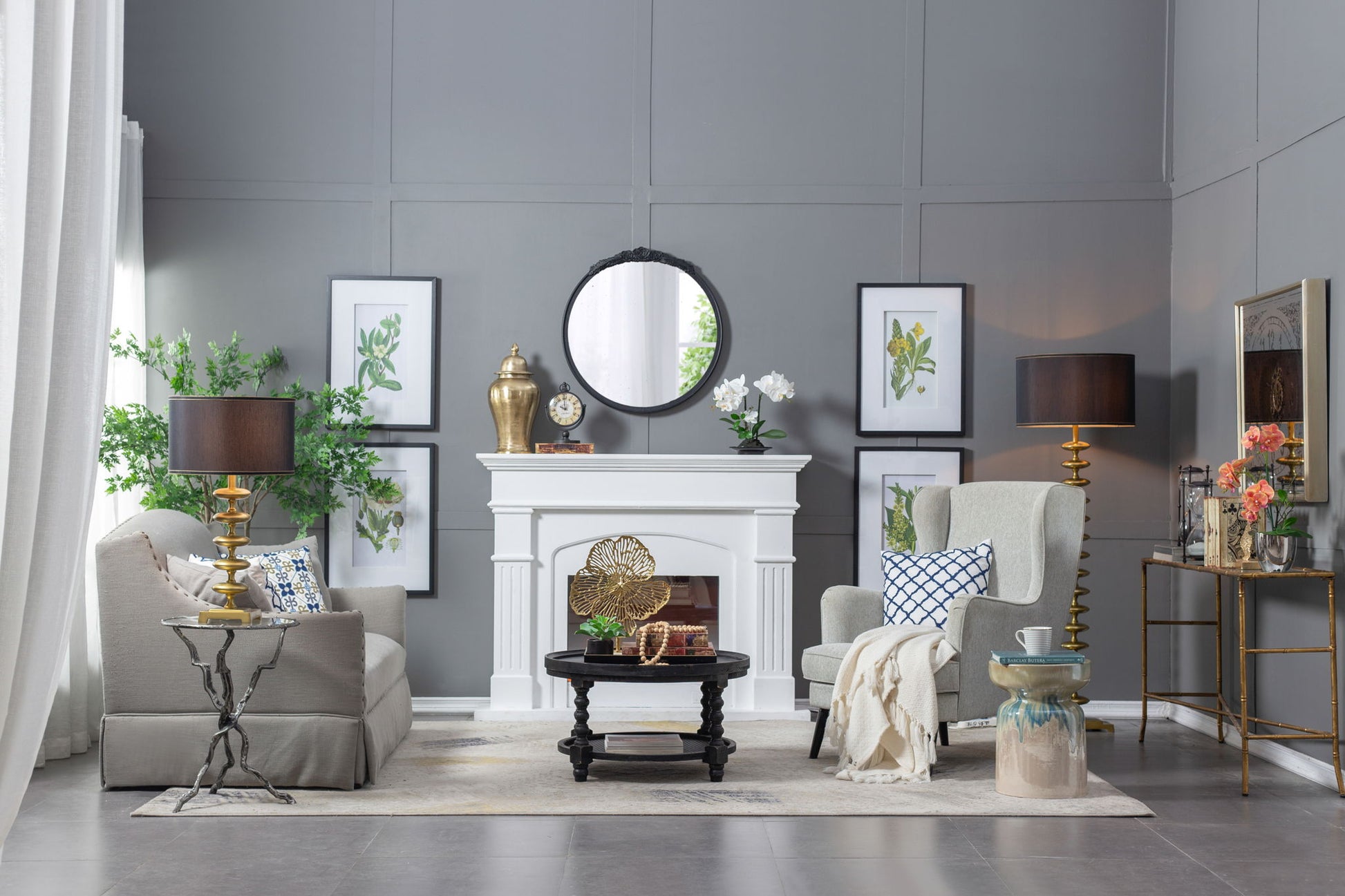 A modern living room with gray walls, a white fireplace, beige sofa, patterned armchair, black coffee table, gold lamps, indoor plants, botanical prints, and the Round Hand Carved Rose Antique Mirror Frame as a stunning wall mirror accent.