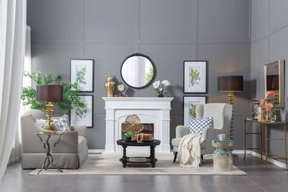 A modern living room with gray walls, a white fireplace, beige sofa, patterned armchair, black coffee table, gold lamps, indoor plants, botanical prints, and the Round Hand Carved Rose Antique Mirror Frame as a stunning wall mirror accent.