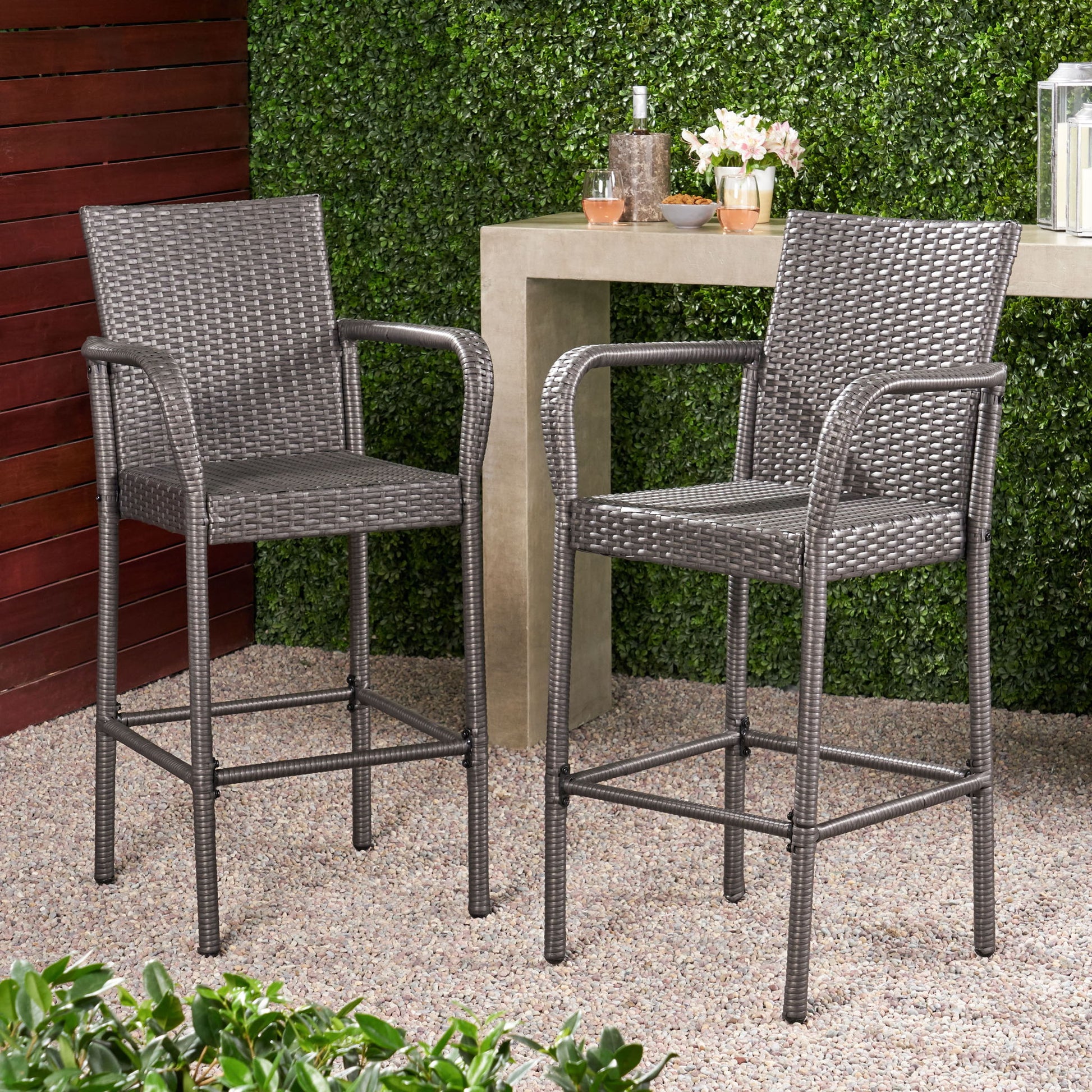 Two Outdoor Wicker Barstools with rattan and iron frames, featuring armrests, sit on a gravel patio beside a bar-height table, surrounded by green hedges and potted plants.