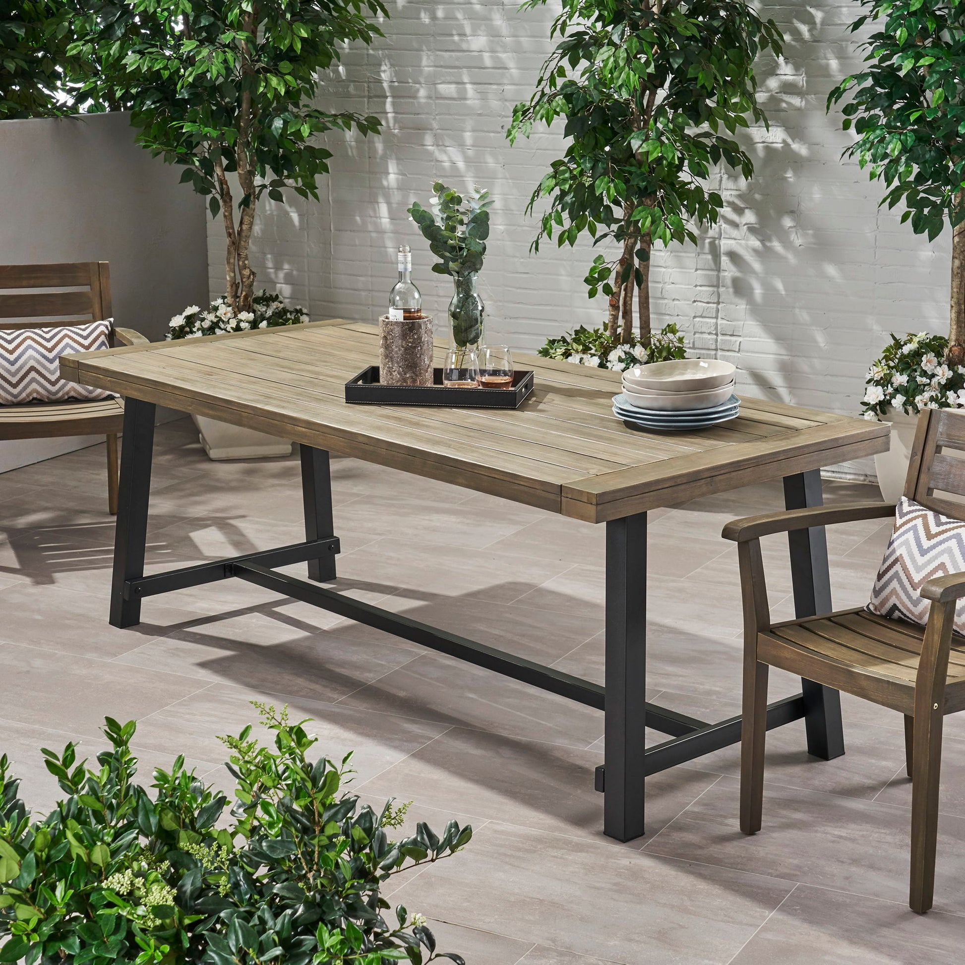 A gray Outdoor Dining Table with rustic metal legs is set with a tray, glass bottles, glasses, and stacked plates. Wooden chairs and green plants surround the table on a tiled patio.