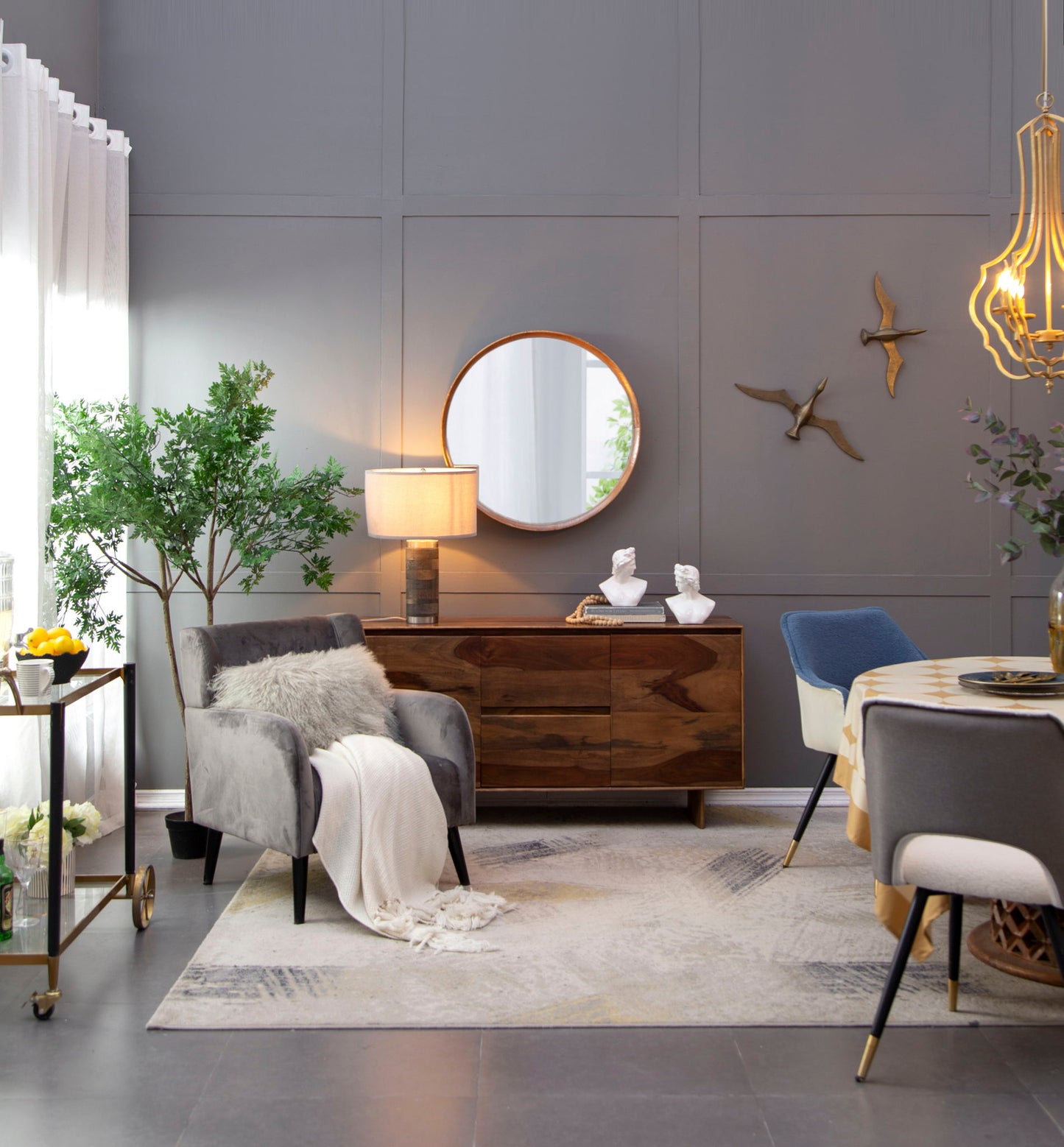 A modern living room with gray walls features the Round Wood Mirror wall mounted as decor, wall art, a wooden sideboard, an armchair with a fur pillow and throw, a round dining table, gold chandelier, green plants, and sheer window curtains.