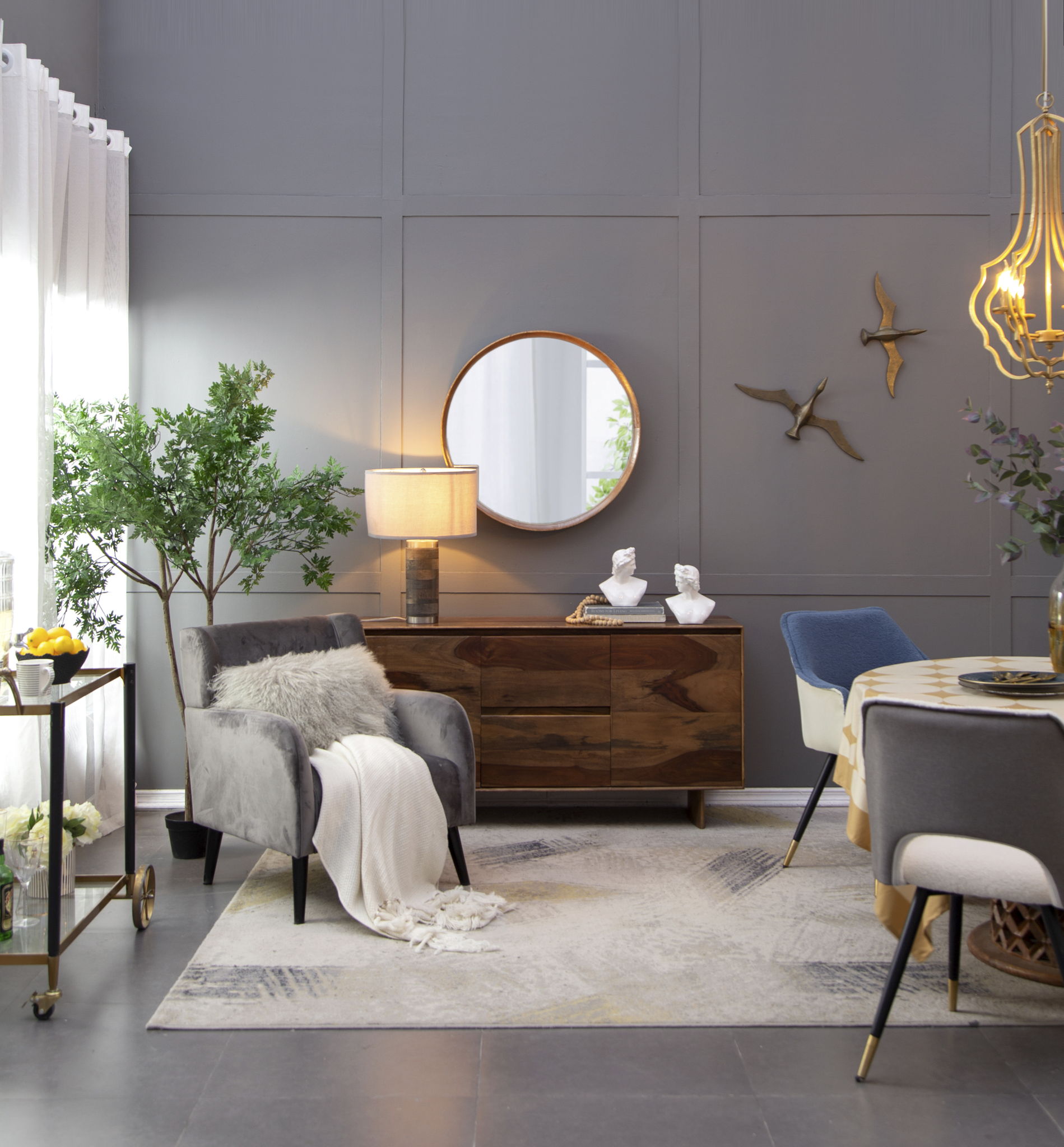 A modern living room with gray walls features the Round Wood Mirror wall mounted as decor, wall art, a wooden sideboard, an armchair with a fur pillow and throw, a round dining table, gold chandelier, green plants, and sheer window curtains.