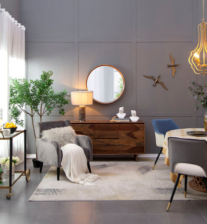 A modern living room with gray walls features the Round Wood Mirror wall mounted as decor, wall art, a wooden sideboard, an armchair with a fur pillow and throw, a round dining table, gold chandelier, green plants, and sheer window curtains.
