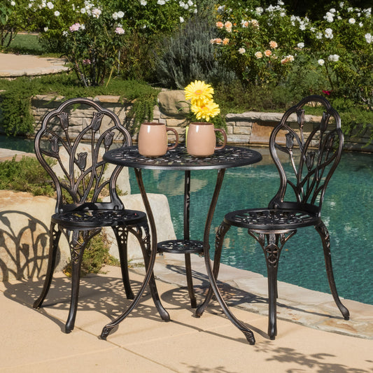 The Nassau - Cast 3 Piece Bistro Set, featuring two ornate black metal chairs and a matching round table, sits by the pool with beige mugs and a vase of yellow flowers on top, surrounded by lush green plants and blooming flowers.