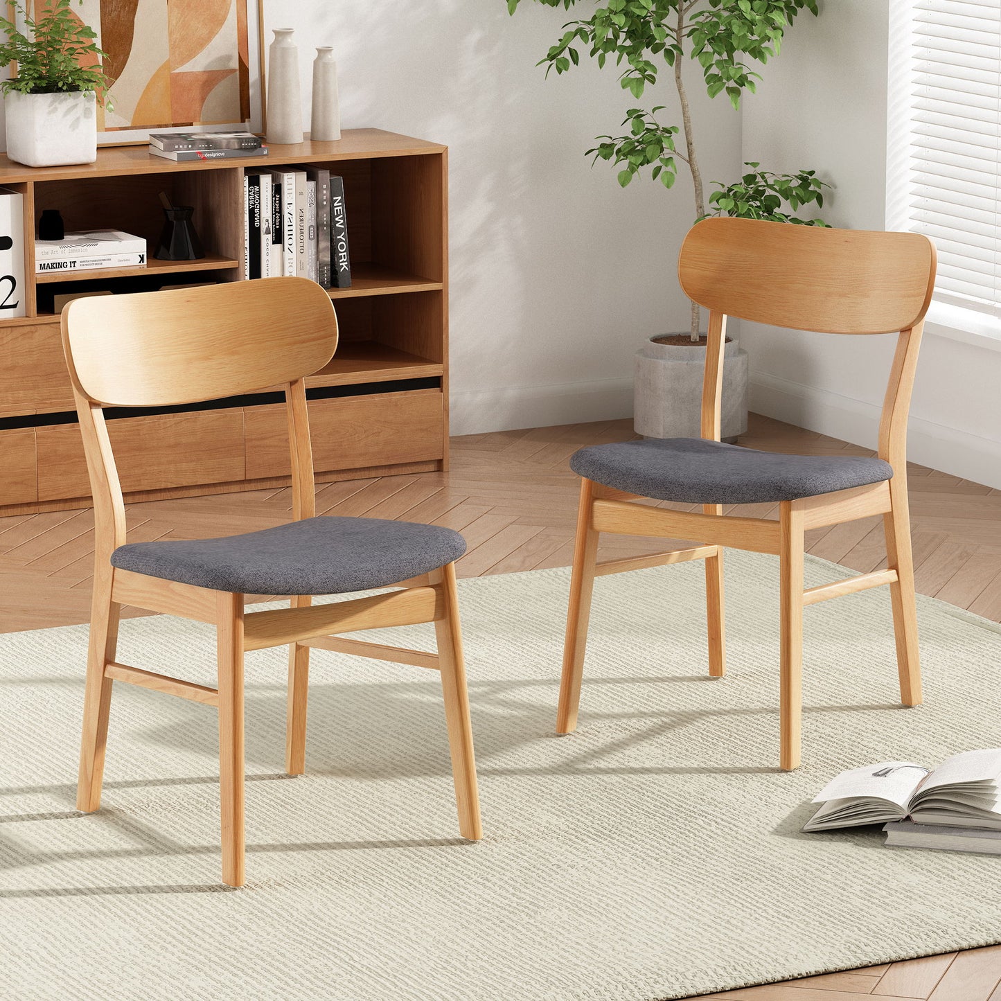 The Brilliantine Dining Chair With Cushion And Wood Frame (Set of 2) in light wood with gray cushions sits on a light rug in a modern living room featuring a wooden bookshelf, books, plants, and sunlight streaming through window blinds.