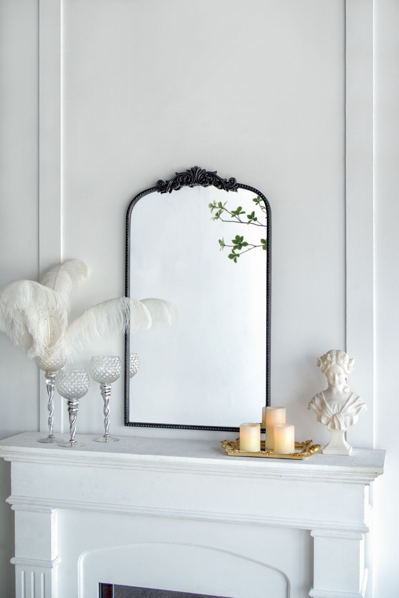A white mantel with decorative feathers in glass holders, three lit candles on a gold tray, a small white bust, and the Arch Mirror, Baroque Inspired Wall Decor reflecting green leaves—all set against a white wall.