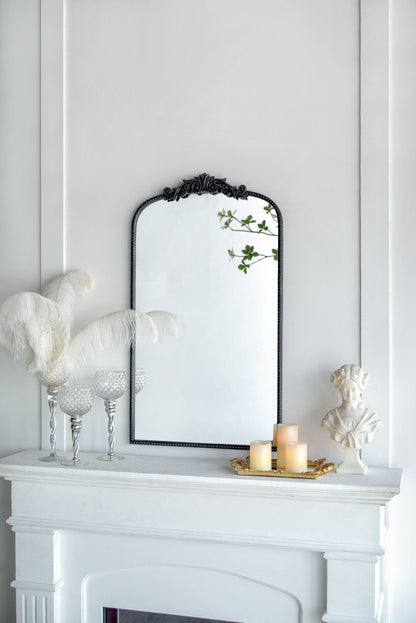 A white mantel with decorative feathers in glass holders, three lit candles on a gold tray, a small white bust, and the Arch Mirror, Baroque Inspired Wall Decor reflecting green leaves—all set against a white wall.