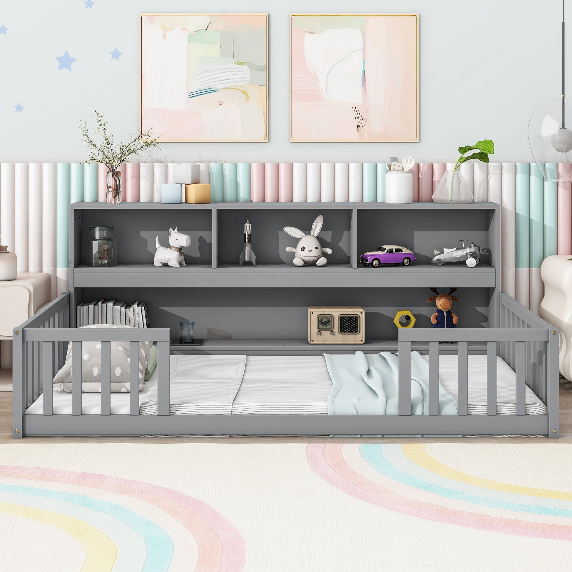 A tidy children’s room features the Floor Bed With Bedside Bookcase, Shelves, Guardrails in gray, filled with toys and books. Soft pastel wall art and a rainbow rug create a playful and cozy atmosphere.