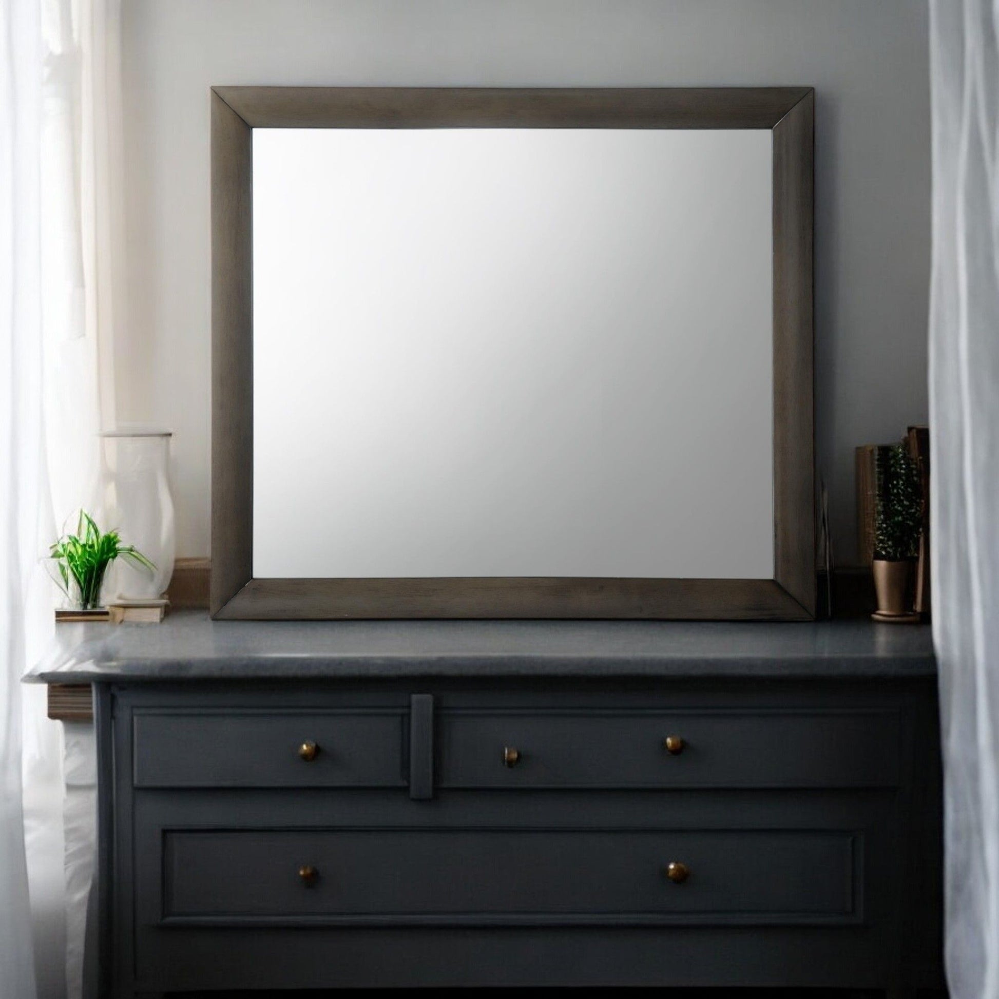 The Ireland Contemporary Design Mirror, a large rectangle with a wooden frame, is mounted above a dark gray dresser featuring brass knobs. A small plant and vase adorn it, while curtains on the left window let in soft light.