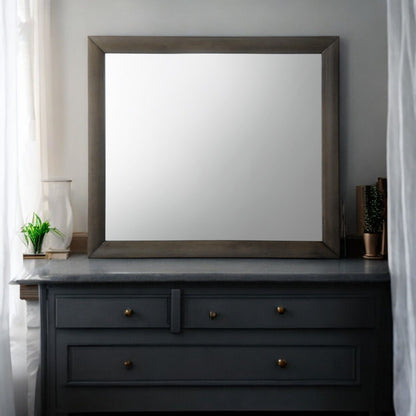 The Ireland Contemporary Design Mirror, a large rectangle with a wooden frame, is mounted above a dark gray dresser featuring brass knobs. A small plant and vase adorn it, while curtains on the left window let in soft light.