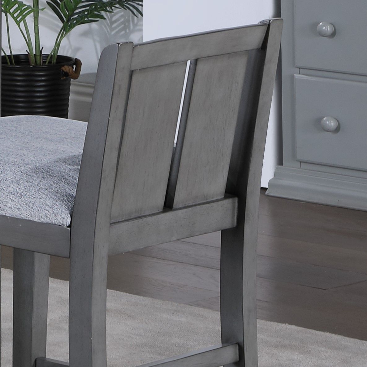 A close-up of the Graham 20" Upholstered Seat Counter Height Chair (Set of 2) in gray, featuring padded seats, placed on a light rug beside a matching gray dresser and a potted plant in the background.